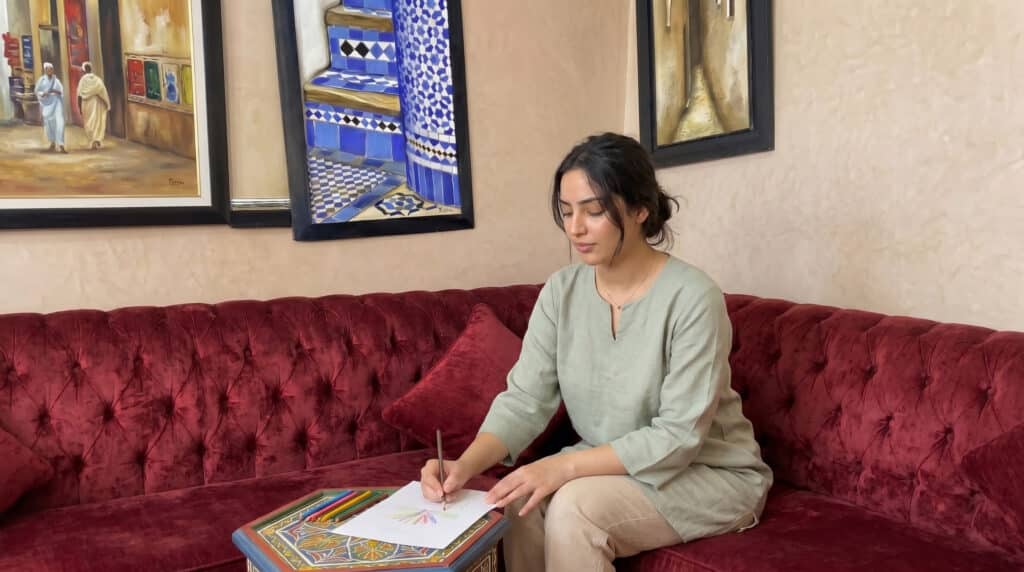 Jeune femme marocaine assise dans un salon marocain, dessinant calmement pour exprimer ses émotions à travers l’art-thérapie.